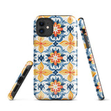 Statement Cases' Mediterranean Bloom for iPhone is showcased, featuring an intricate, colorful pattern. The design includes blue, yellow, and orange floral shapes in a symmetrical arrangement on a light blue background. This durable phone case provides dual-layer protection and is constructed from impact-resistant polycarbonate to safeguard your device.