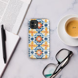 Statement Cases' Mediterranean Bloom for iPhone is showcased, featuring an intricate, colorful pattern. The design includes blue, yellow, and orange floral shapes in a symmetrical arrangement on a light blue background. This durable phone case provides dual-layer protection and is constructed from impact-resistant polycarbonate to safeguard your device.