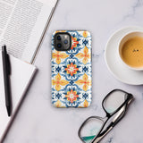 Statement Cases' Mediterranean Bloom for iPhone is showcased, featuring an intricate, colorful pattern. The design includes blue, yellow, and orange floral shapes in a symmetrical arrangement on a light blue background. This durable phone case provides dual-layer protection and is constructed from impact-resistant polycarbonate to safeguard your device.