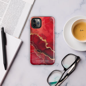 A smartphone in a protective Scarlet Marble for iPhone case by Statement Cases with a red and gold marbled abstract design. The case has "Statement Cases" written at the bottom. The iPhone 15 Pro Max features three camera lenses and a flash on the top left corner.