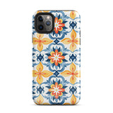 Statement Cases' Mediterranean Bloom for iPhone is showcased, featuring an intricate, colorful pattern. The design includes blue, yellow, and orange floral shapes in a symmetrical arrangement on a light blue background. This durable phone case provides dual-layer protection and is constructed from impact-resistant polycarbonate to safeguard your device.