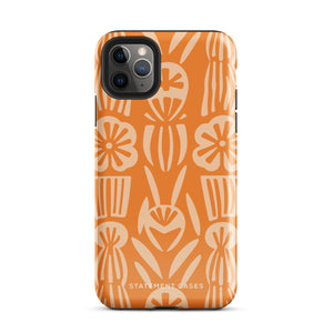 A Savannah Ardiente for iPhone from Statement Cases, featuring a dual-layered phone case in orange with a floral pattern showcasing cream-colored flowers, leaves, and geometric shapes. This impact-resistant polycarbonate case displays "STATEMENT CASES" at the bottom and reveals the camera lenses at the top left corner.