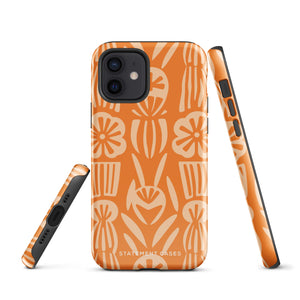 A Savannah Ardiente for iPhone from Statement Cases, featuring a dual-layered phone case in orange with a floral pattern showcasing cream-colored flowers, leaves, and geometric shapes. This impact-resistant polycarbonate case displays "STATEMENT CASES" at the bottom and reveals the camera lenses at the top left corner.