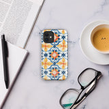 Statement Cases' Mediterranean Bloom for iPhone is showcased, featuring an intricate, colorful pattern. The design includes blue, yellow, and orange floral shapes in a symmetrical arrangement on a light blue background. This durable phone case provides dual-layer protection and is constructed from impact-resistant polycarbonate to safeguard your device.