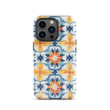 Statement Cases' Mediterranean Bloom for iPhone is showcased, featuring an intricate, colorful pattern. The design includes blue, yellow, and orange floral shapes in a symmetrical arrangement on a light blue background. This durable phone case provides dual-layer protection and is constructed from impact-resistant polycarbonate to safeguard your device.