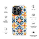 Statement Cases' Mediterranean Bloom for iPhone is showcased, featuring an intricate, colorful pattern. The design includes blue, yellow, and orange floral shapes in a symmetrical arrangement on a light blue background. This durable phone case provides dual-layer protection and is constructed from impact-resistant polycarbonate to safeguard your device.