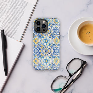 The Blue Mirage for iPhone by Statement Cases features a dual-layered, impact-resistant polycarbonate exterior with a TPU inner liner. Inspired by traditional Portuguese tiles, the blue and yellow patterned case boasts an intricate design of floral and geometric motifs. With its elegant and colorful appearance, the phone's camera is visible at the top left of the case.