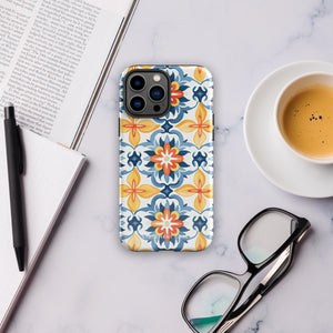 Statement Cases' Mediterranean Bloom for iPhone is showcased, featuring an intricate, colorful pattern. The design includes blue, yellow, and orange floral shapes in a symmetrical arrangement on a light blue background. This durable phone case provides dual-layer protection and is constructed from impact-resistant polycarbonate to safeguard your device.