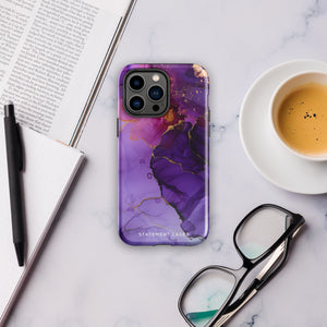 A Statement Cases Golden Orchid Marble for iPhone with a case featuring an abstract design of swirling purple, pink, and gold hues over a white background. The marbled patterns with metallic accents provide dual-layer protection and are impact-resistant. The top of the case has cutouts for the phone's camera lenses and is induction charging compatible.