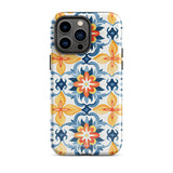 Statement Cases' Mediterranean Bloom for iPhone is showcased, featuring an intricate, colorful pattern. The design includes blue, yellow, and orange floral shapes in a symmetrical arrangement on a light blue background. This durable phone case provides dual-layer protection and is constructed from impact-resistant polycarbonate to safeguard your device.