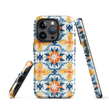 Statement Cases' Mediterranean Bloom for iPhone is showcased, featuring an intricate, colorful pattern. The design includes blue, yellow, and orange floral shapes in a symmetrical arrangement on a light blue background. This durable phone case provides dual-layer protection and is constructed from impact-resistant polycarbonate to safeguard your device.