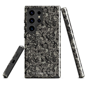 A phone case with a protective dual-layer design featuring an intricate black floral lace pattern. The Omerta Floral for Samsung has four camera cutouts and the brand name "Statement Cases" appears at the bottom center. Impact-resistant and chic, it stands against a plain white background.