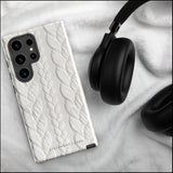 A stylish, impact-resistant phone case with a textured white braided design, covering the back of a smartphone. The dual-layer design features cutouts for the camera and buttons, and "Cozy Knit Bliss for Samsung" by Statement Cases is embossed at the bottom.