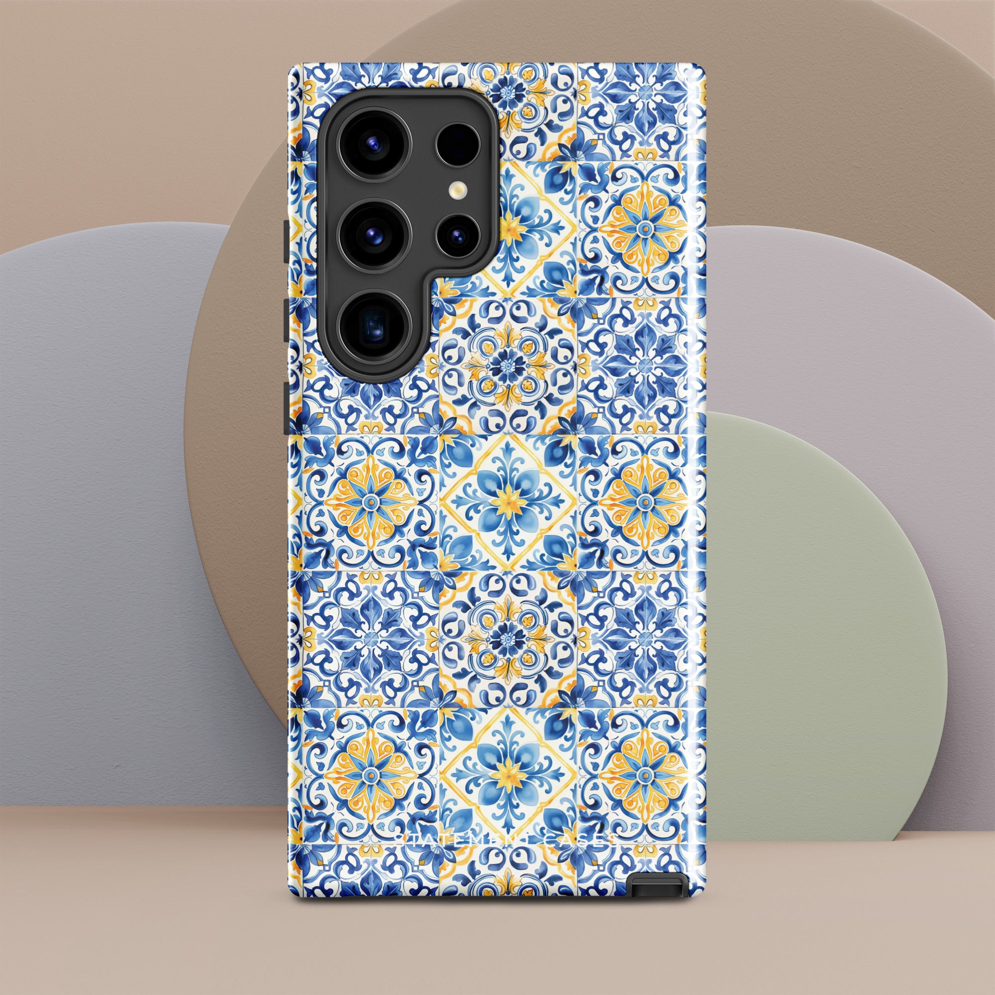 The Blue Mirage for Samsung by Statement Cases is a durable phone case showcasing a detailed blue and yellow tile pattern. The design includes symmetrical floral and geometric motifs reminiscent of classic ceramic tiles. Its impact-resistant dual-layer construction is precisely crafted to fit around the phone's camera and buttons seamlessly.